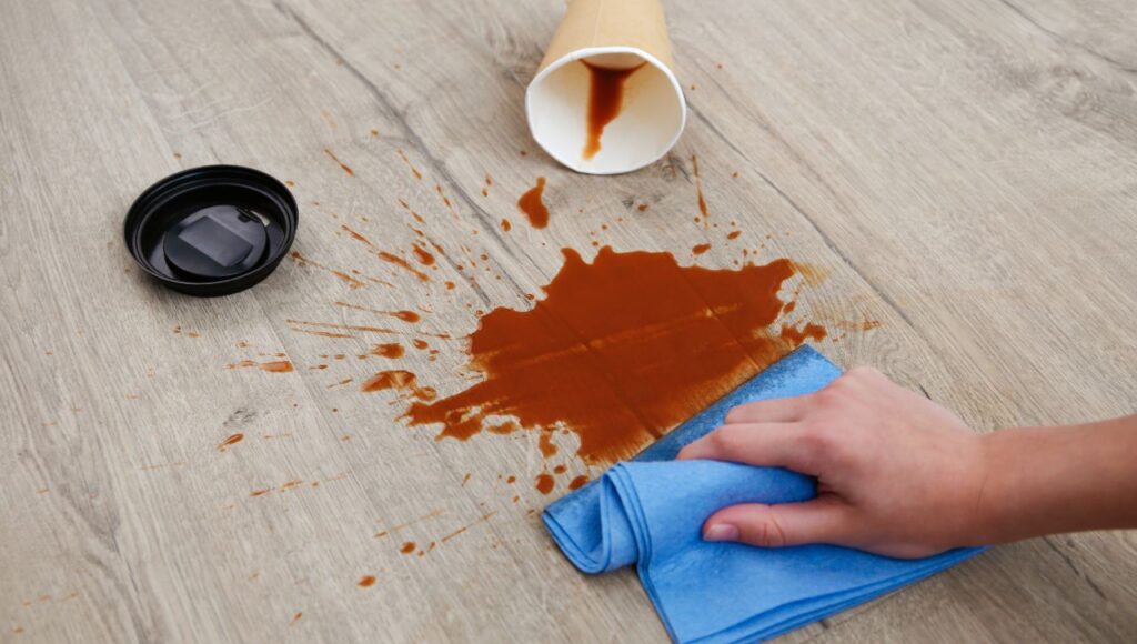 A hand cleaning a coffee spill on a durable laminate floor installed by Orange Cat Flooring, showcasing the flooring's stain-resistant surface.