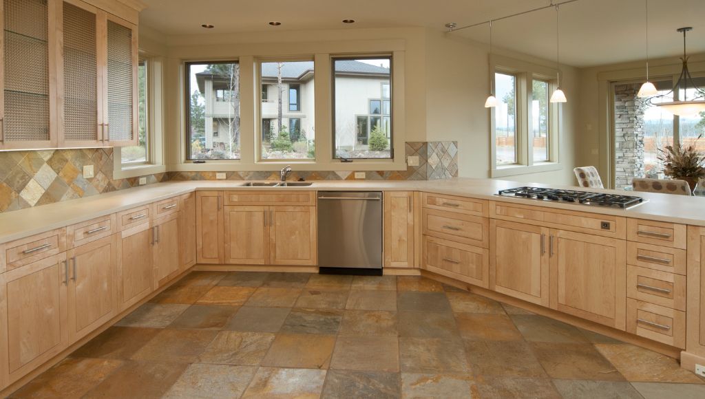 Modern kitchen with natural stone tile flooring and backsplash, installed by Orange Cat Flooring, featuring light wood cabinetry and large windows.